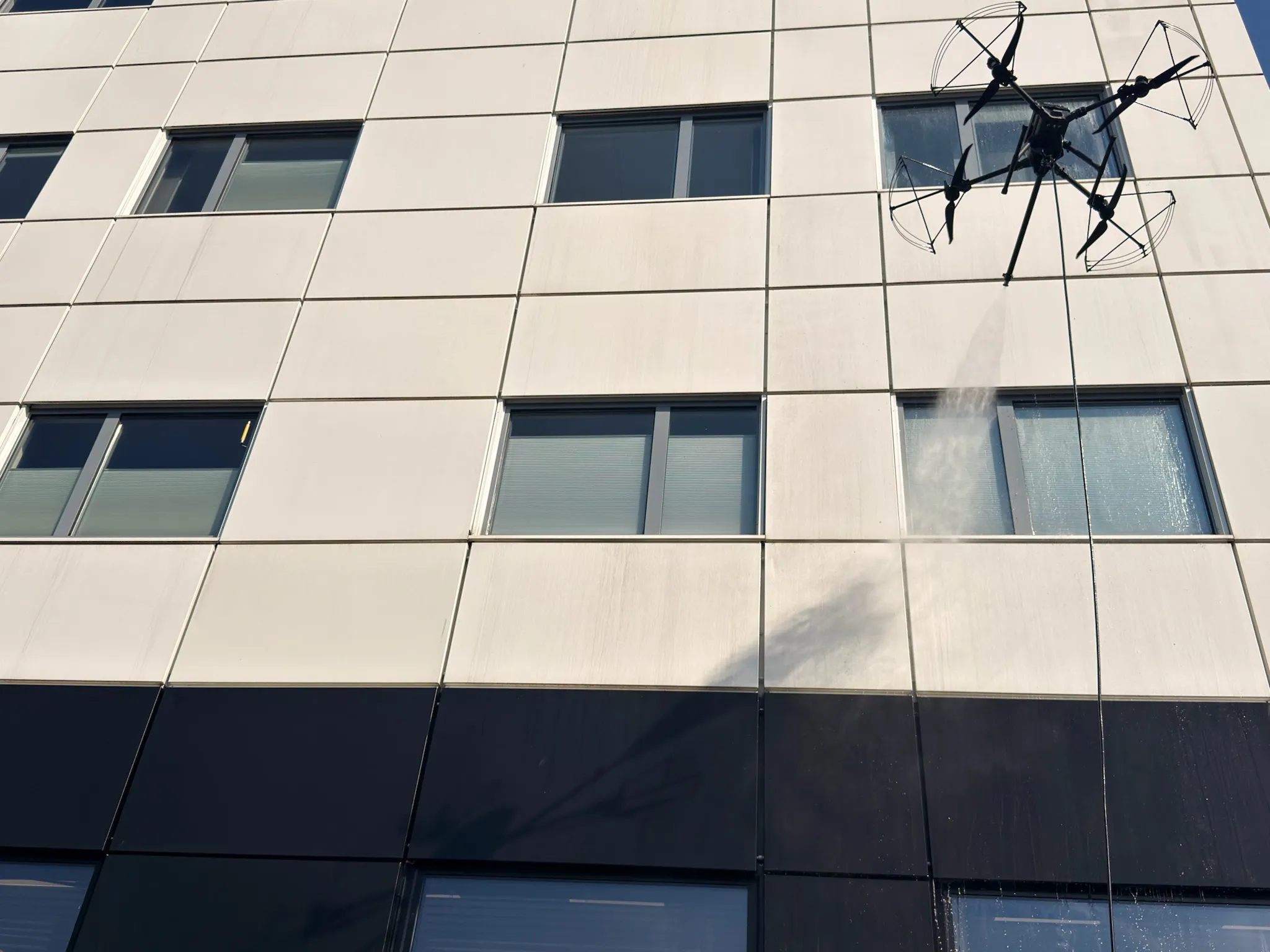 En vaskedrone svever inntil fasaden på et bygg og sprøyter vann mot betongplatene, sett fra bakken